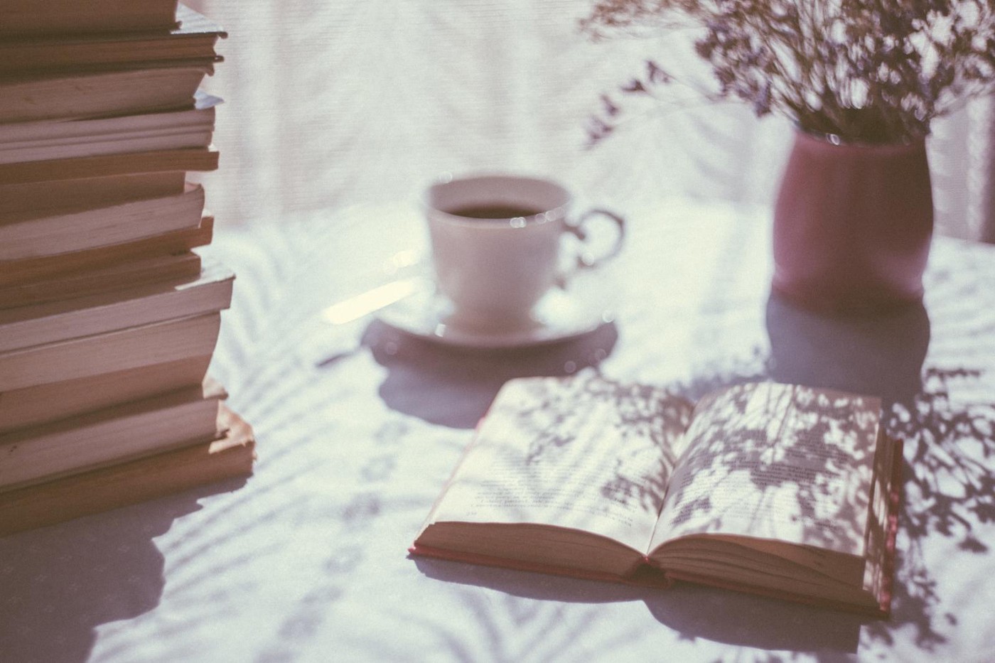A coffee table next to a bright window, on top of which sits a stack of books, a cup of tea, an open book, and a vase.