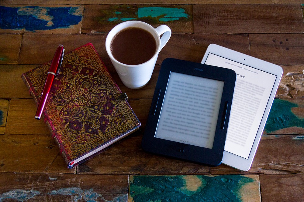 An ebook reader, iPad, a notebook and pen, and a cup of hot chocolate all on a rough wooden table.